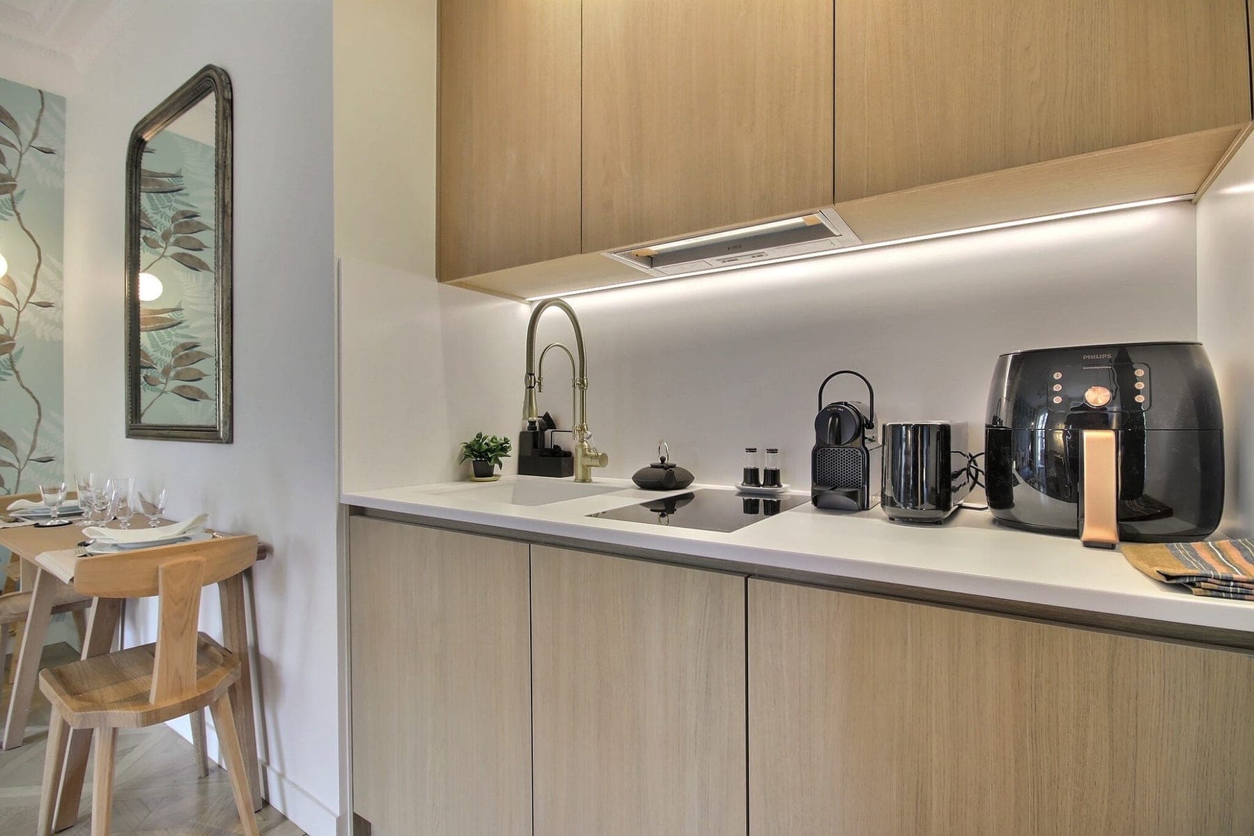 Cuisine linéaire en bois clair d’un meublé parisien, plan de travail blanc et éclairage sous meubles
